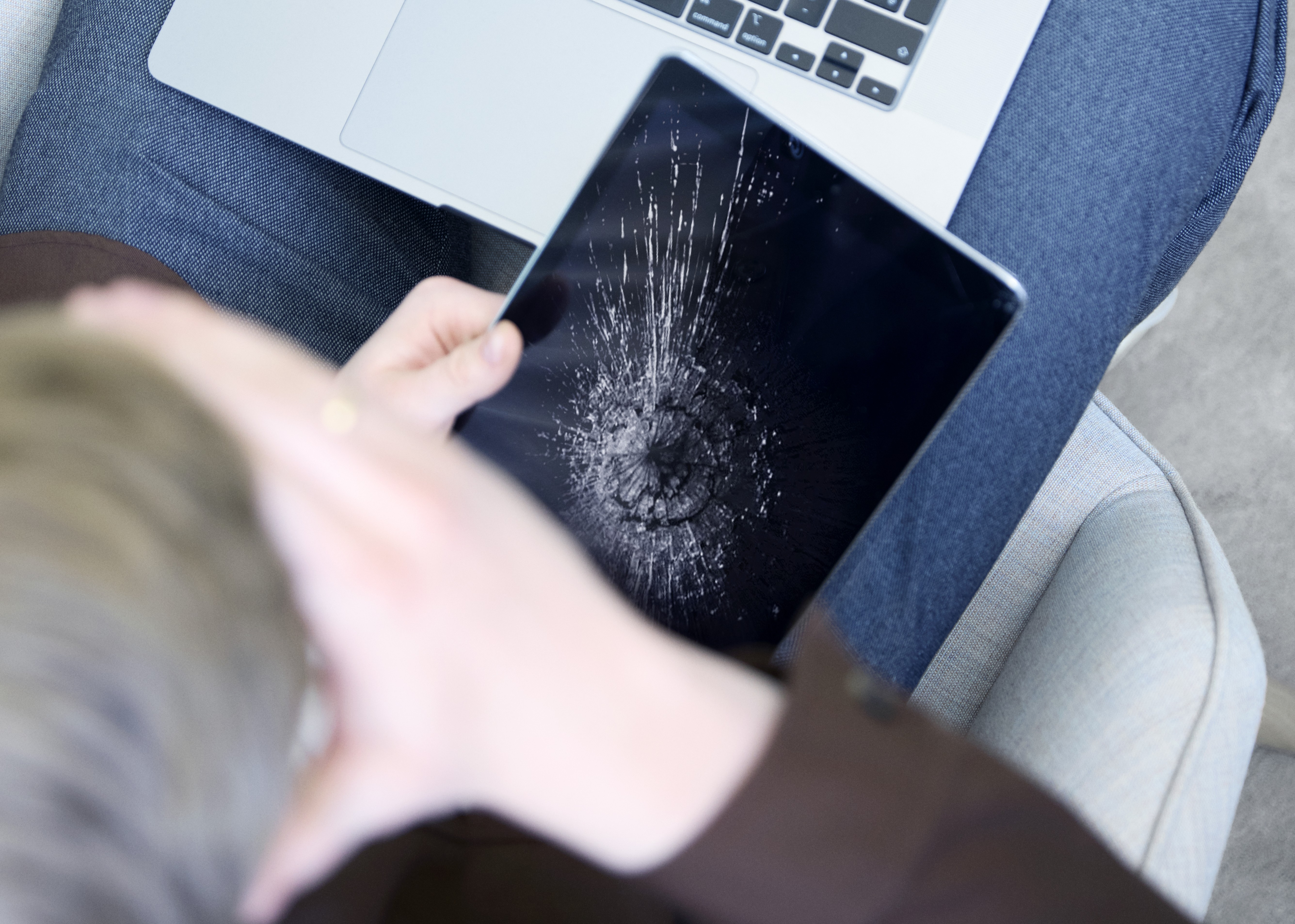 En person holder en sort tablet med knust skærm over et skød med en laptop i baggrunden.